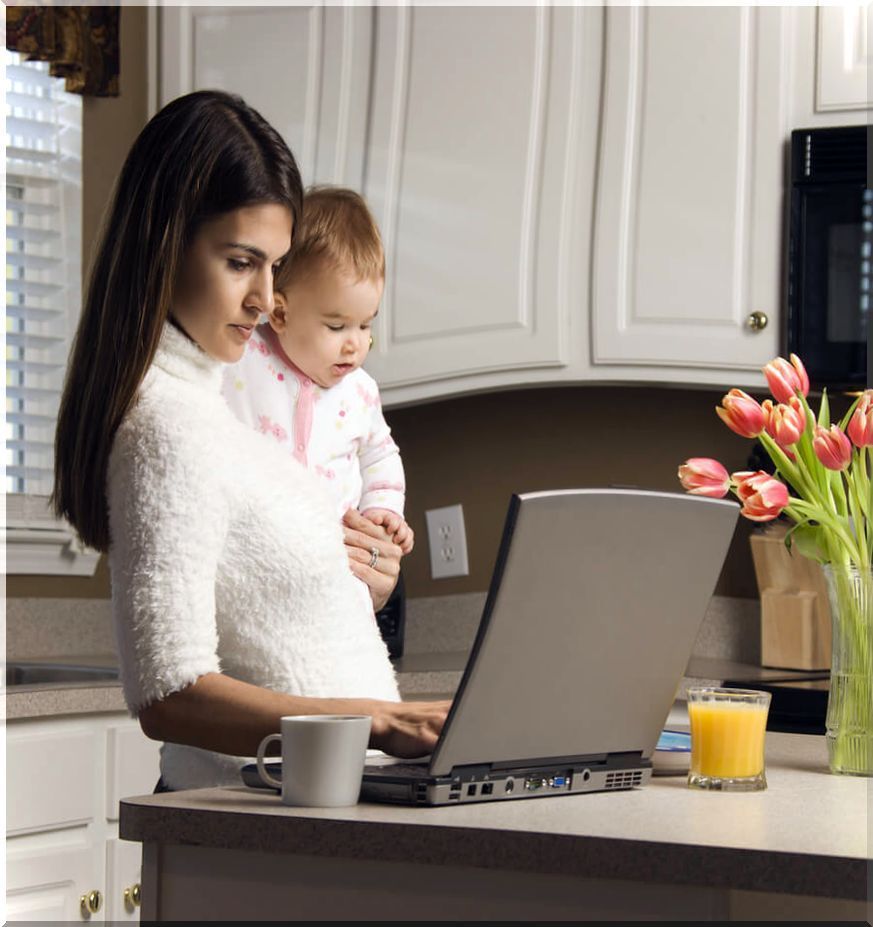 Mother holding her baby while typing on laptop in kitchen.